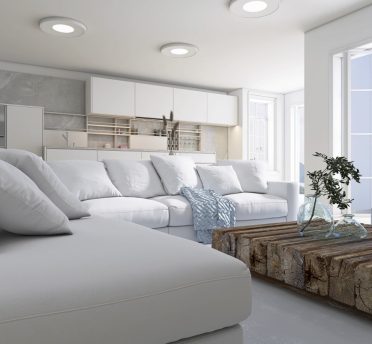 Un soggiorno moderno con un divano componibile bianco e cuscini. Un tavolino da caffè rustico in legno espone vasi di vetro. Lo spazio è luminoso, con grandi finestre che lasciano entrare la luce naturale. La zona cucina aperta è sullo sfondo con mobili bianchi.