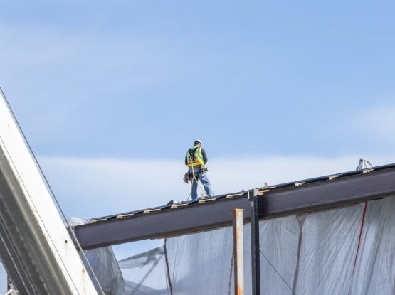 Un operaio edile con un giubbotto di sicurezza e un casco Cila ROma è in piedi su una trave di acciaio nel cantiere. L'operaio è sollevato, incorniciato da un cielo parzialmente nuvoloso. Impalcature e teli di plastica si estendono sotto di lui.
