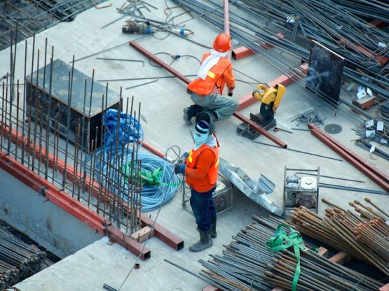 Due operai edili in un cantiere di Cila Roma. Uno è inginocchiato mentre usa una sega su travi metalliche, e l'altro è in piedi lì vicino, a sorvegliare. Il cantiere è pieno di barre, cavi e barre di rinforzo su una soletta di cemento. Entrambi indossano equipaggiamento di sicurezza e caschi.