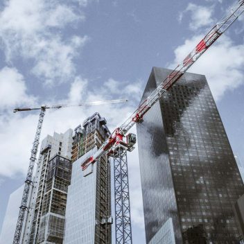 Due moderni grattacieli con facciate in vetro riflettente in costruzione. Due grandi gru sono visibili contro un cielo azzurro con nuvole sparse, che assistono al processo di costruzione.